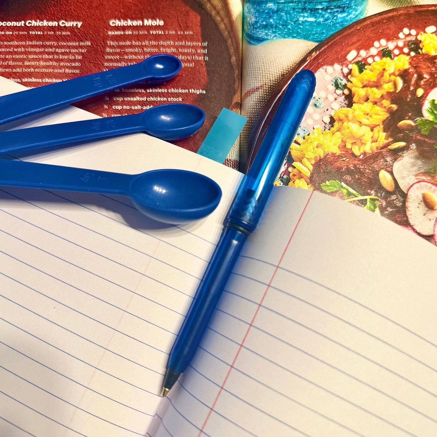 Blue measuring spoons and pen on a recipe book with a food photograph