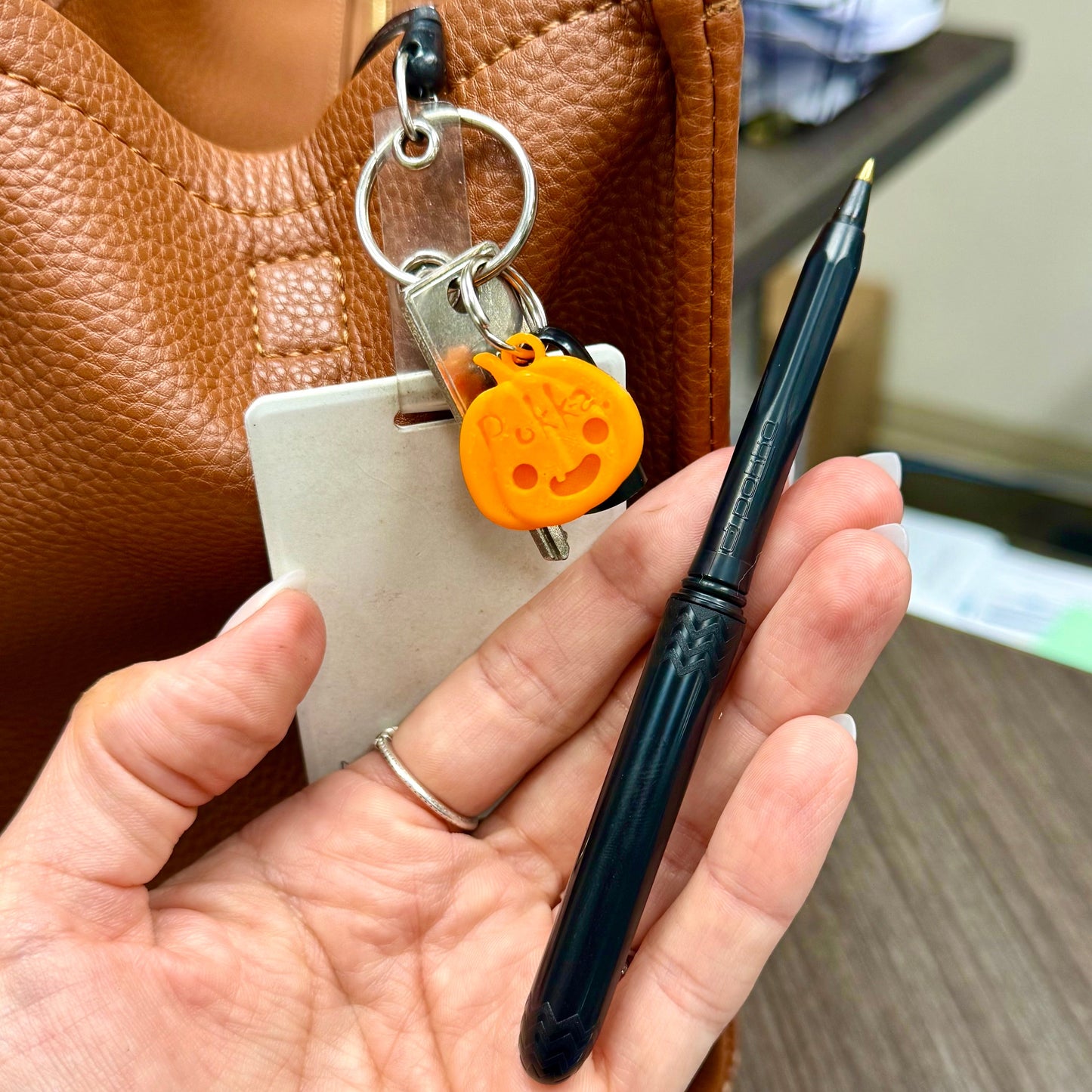 Hand holding a pen with a keychain and small orange object, next to a brown leather bag.
