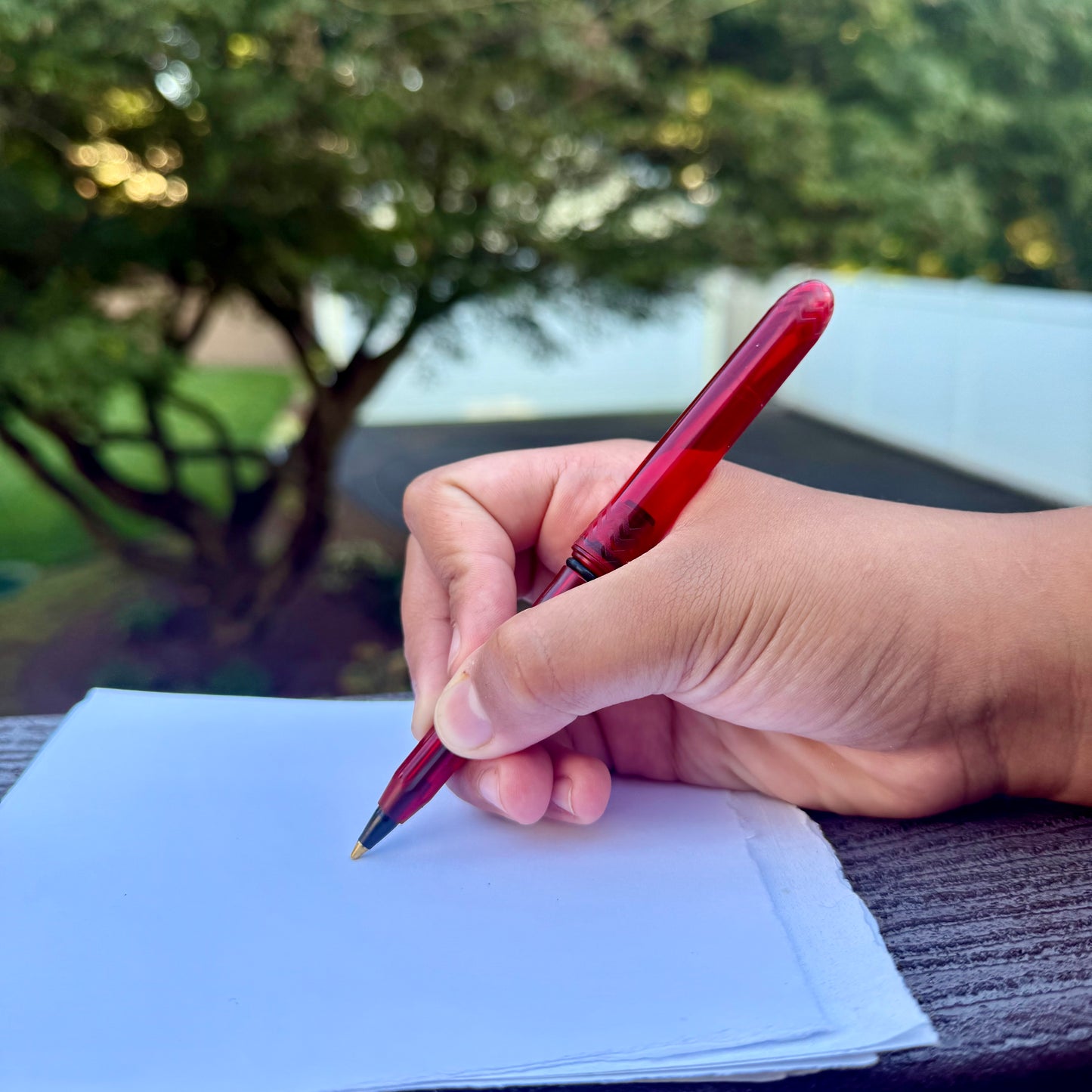 Hand holding a red pen over a piece of paper with a blurred outdoor background