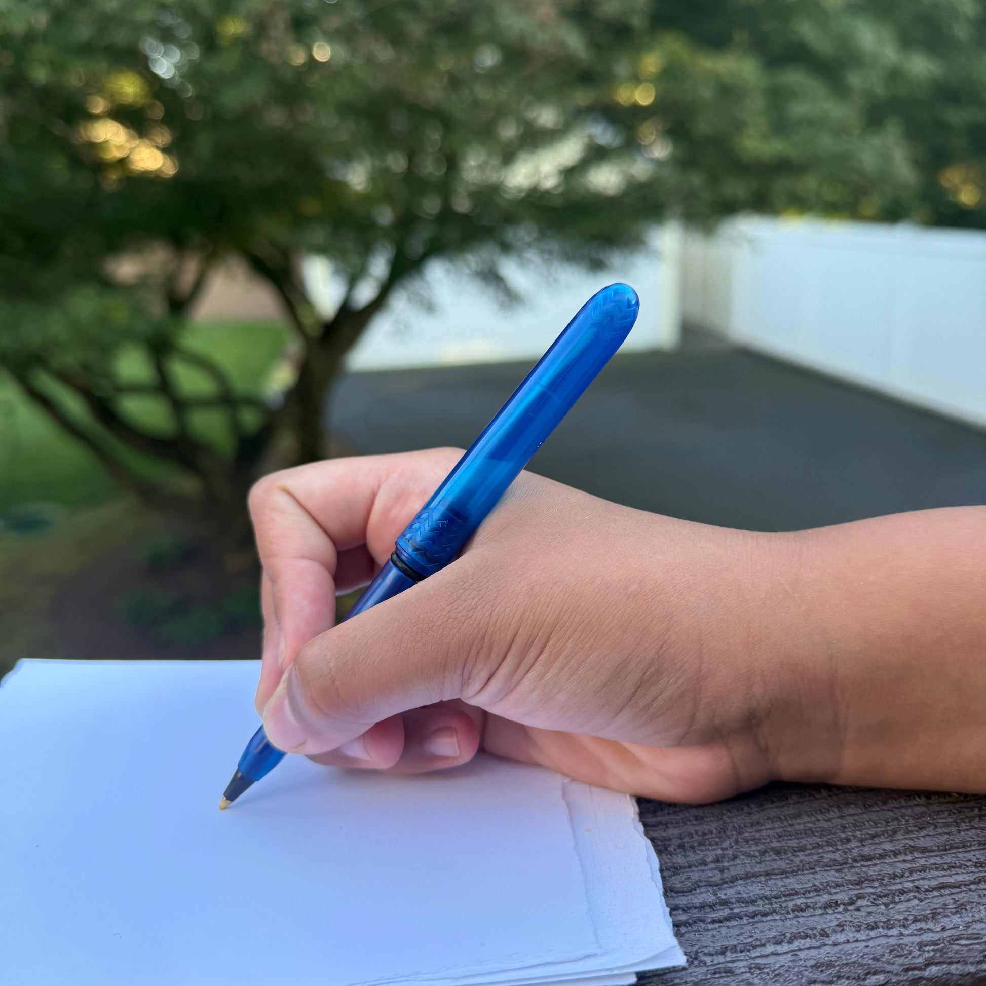 Hand holding a blue pen over a piece of paper with a blurred outdoor background