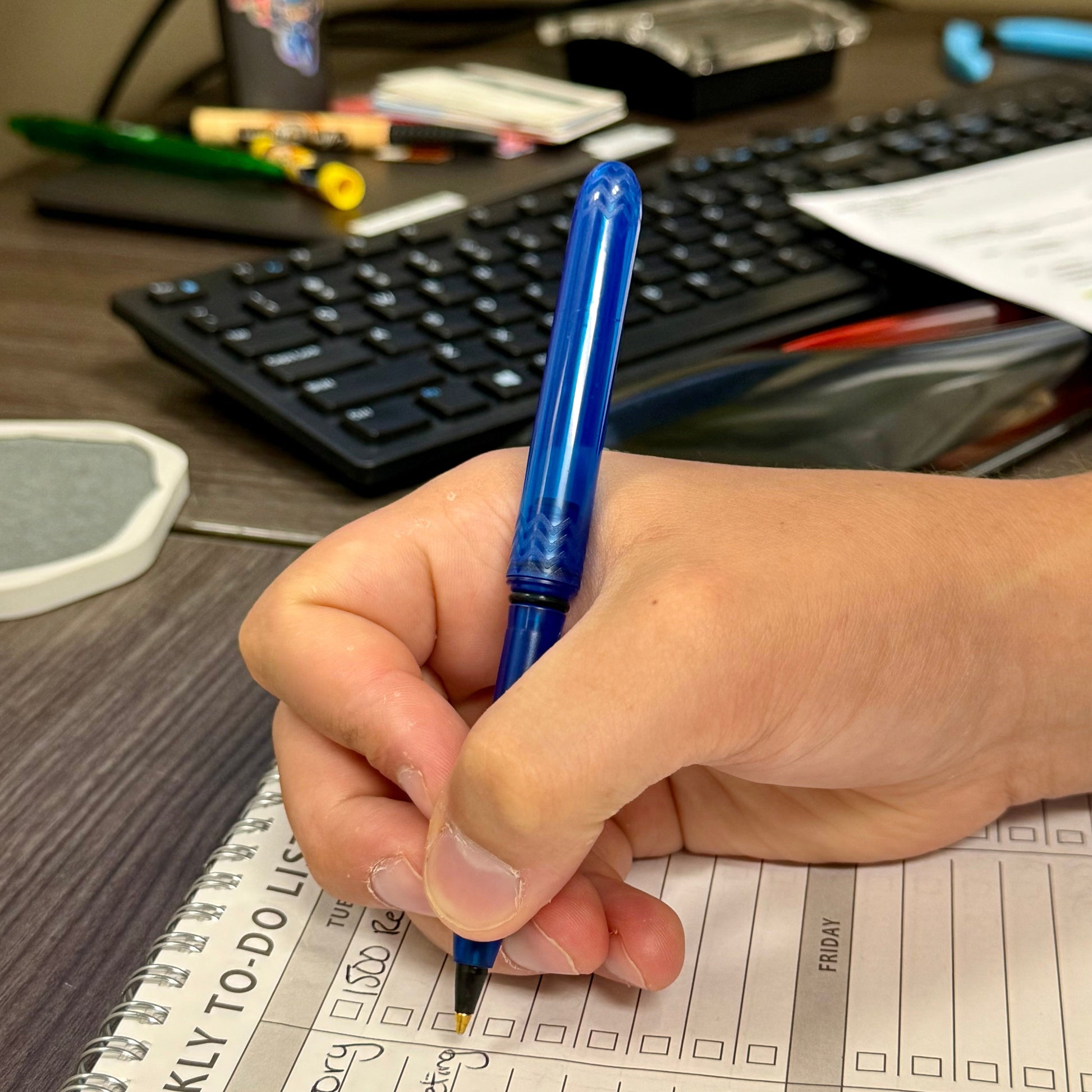 Hand holding a blue pen over a weekly to-do list on a desk with a keyboard in the background.