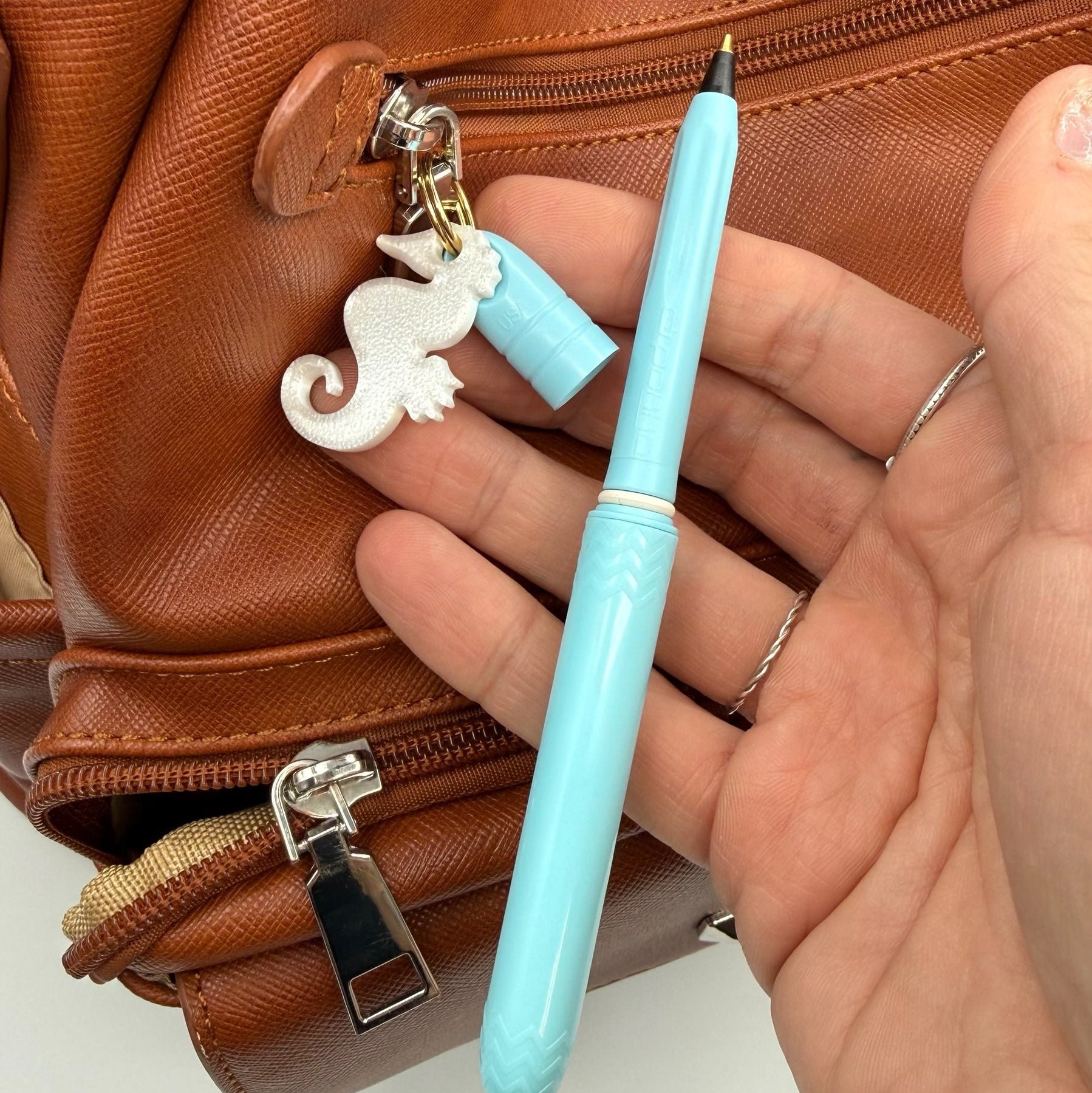 Hand holding a light blue pen with a decorative charm in front of a brown leather bag.